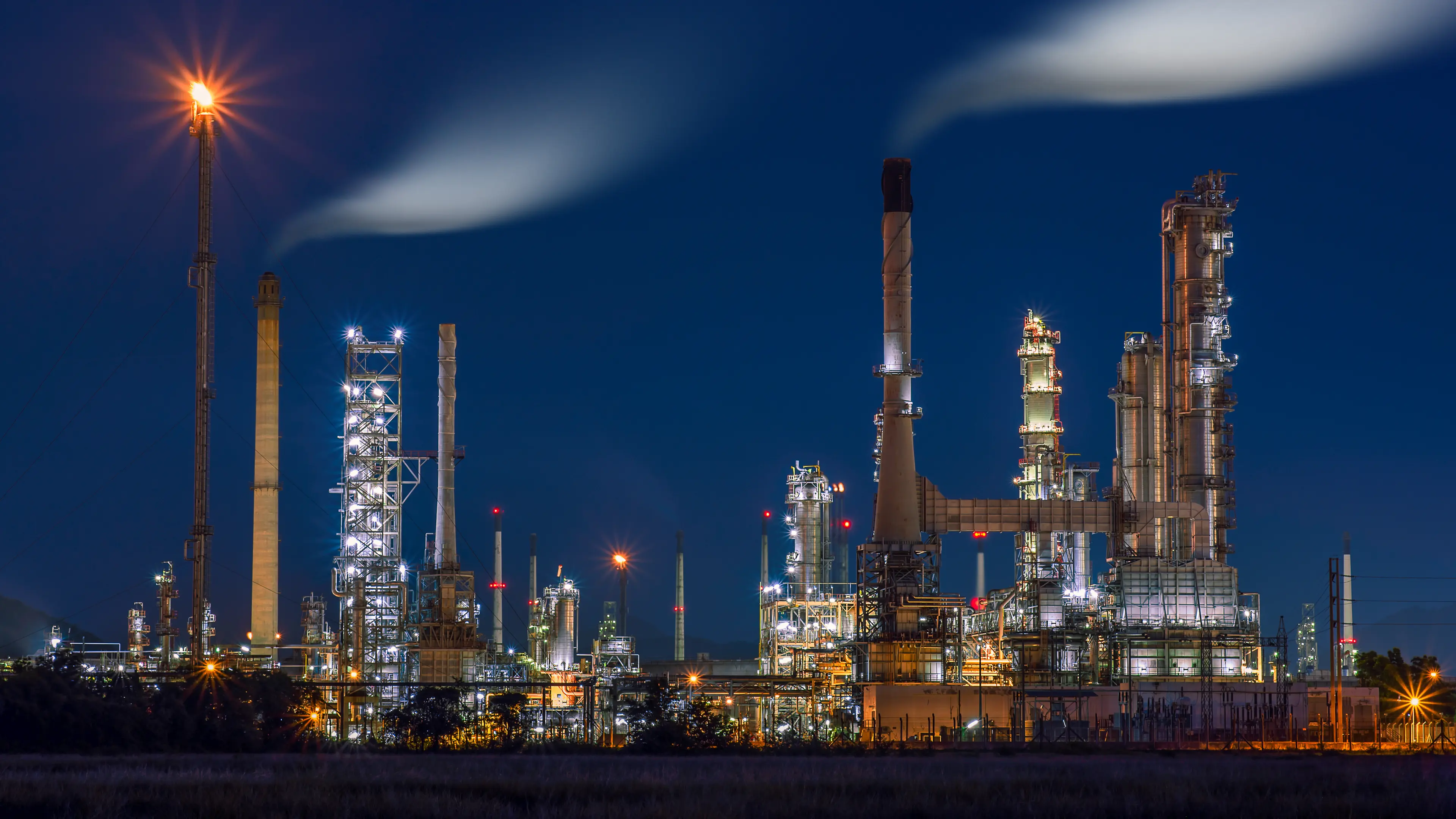 Illuminated oil refinery and energy infrastructure at night, tall chimneys emitting smoke under a dark blue sky.