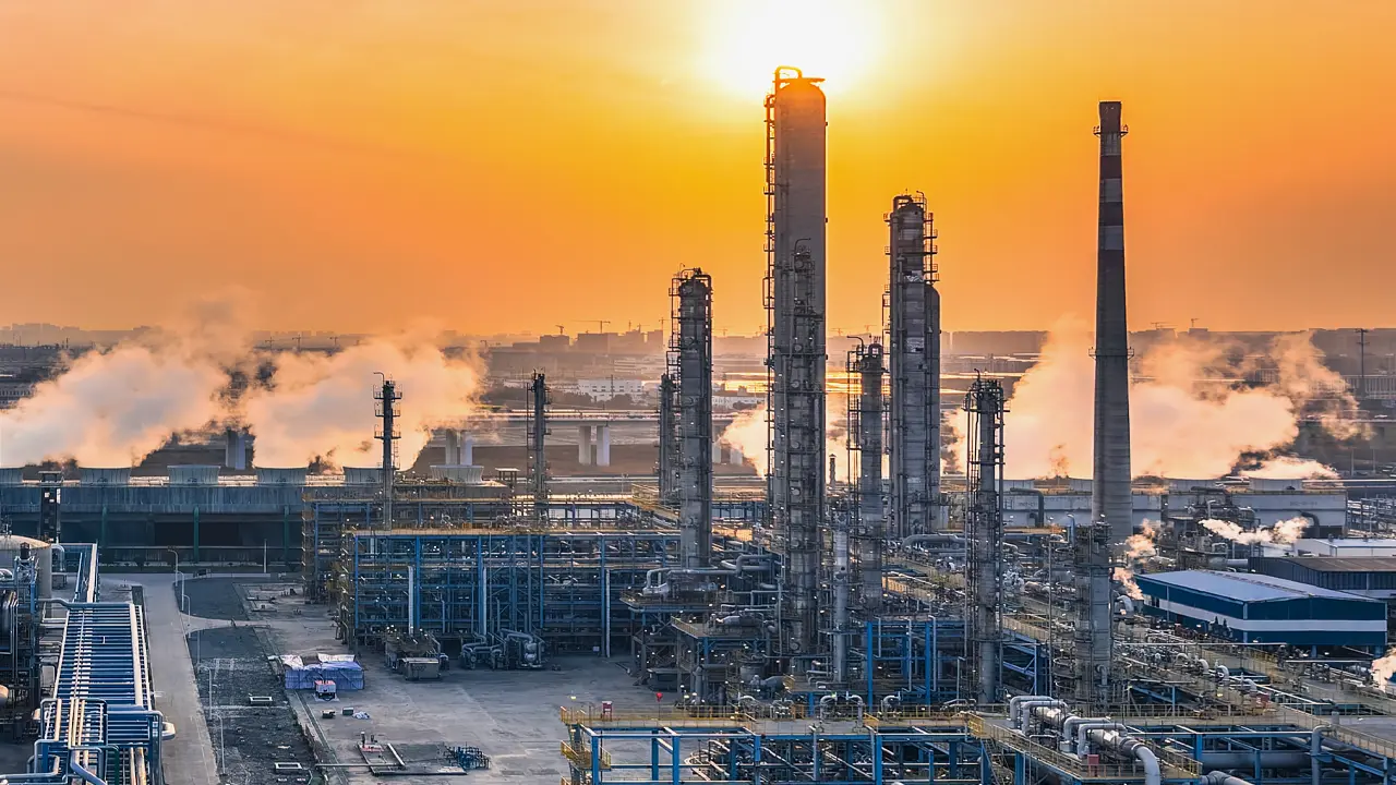 Industrial petrochemical refinery at sunset with tall chimneys emitting smoke, silhouetted against an orange sky.
