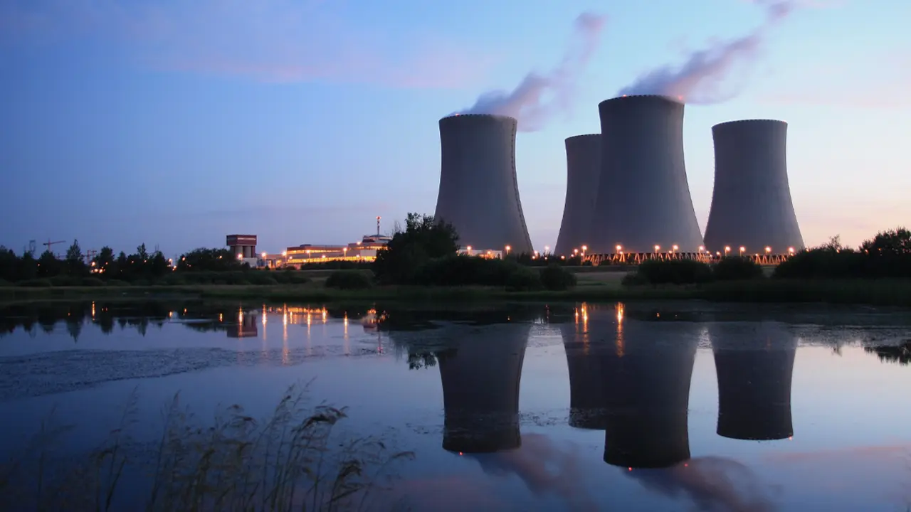 Cooling towers of a power plant emitting steam at twilight, reflecting in water, symbolizing electric generation.