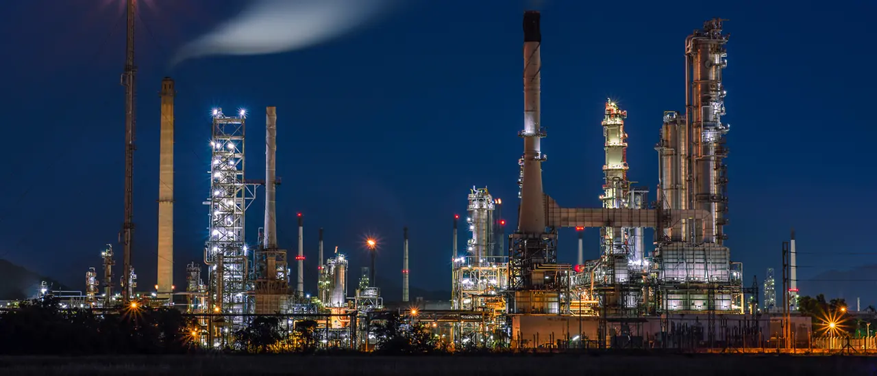 Brightly lit industrial refinery at night, with towering chimneys and structures. The deep blue sky contrasts with the glowing lights.
