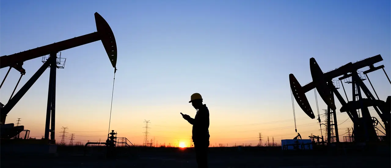 Silhouetted oil pumps and a worker at sunset. The worker, wearing a hard hat, looks at a phone against a vibrant orange and blue sky.