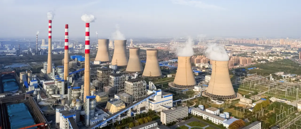 Aerial view of an industrial power plant with cooling towers and chimneys releasing steam near a city skyline.