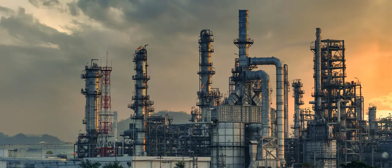 A brightly lit industrial refinery at night, with towering chimneys and intricate metal structures set against a deep blue sky. The scene conveys a sense of industrial activity.