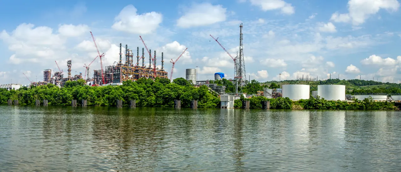 : Ethylene cracker complex with cranes and storage tanks beside a river, separated by greenery under a blue sky.